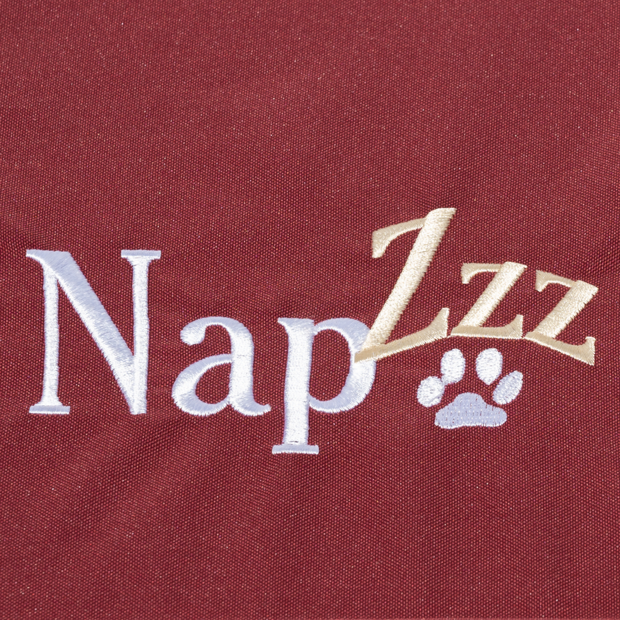 Napzzz benchmatras bordeaux rood in vooraanzicht als detail foto van stof en geborduurde napzzz logo. Gemaakt van waterafstotende stof genaamd oxford. Stevige schuimrubberen matras met een oxford hoes voorzien van ritssluiting. Mooi afgewerkt, stevig, comfortabel matras voor in benches. Met geborduurde napzzz logo als leuk detail
